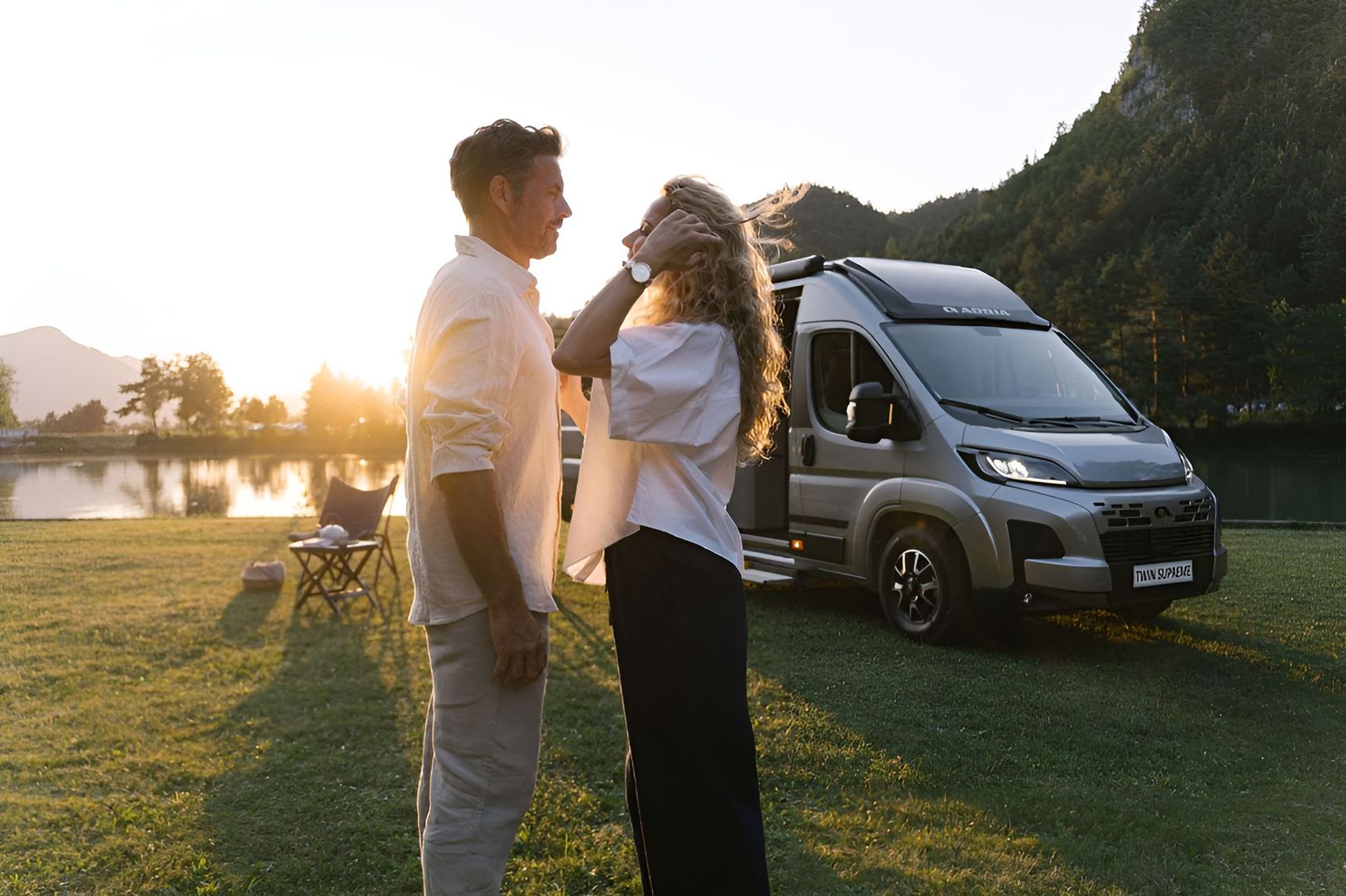 Zwei Personen stehen auf einer Wiese vor einem geparkten Wohnmobil bei Sonnenuntergang mit Bergen und See im Hintergrund